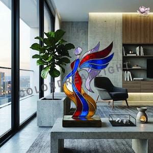 May include: A stained glass angel sculpture with vibrant red, blue, and gold hues. The artwork features a flowing design, mounted on a wooden base. The sculpture is displayed on a modern concrete table, with the text "COLOR DREAM LIGHT" visible.