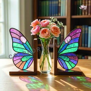Könnte beinhalten: Bücherstützen in Form von Schmetterlingsflügeln aus Buntglas in Lila, Blau und Grün. Die Buchstützen sind aus Holz und halten eine klare Glasvase mit rosa Rosen. Die Buchstützen stehen auf einem Holztisch.