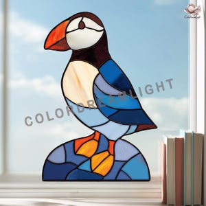Puede incluir: Vidriera de frailecillo con un diseño vibrante. El ave presenta un gran pico naranja, cara blanca y cuerpo azul y negro. El ave está posada sobre una base azul en forma de roca. El texto "COLORDREAMLIGHT" es visible.