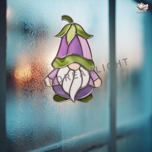 May include: A stained glass gnome decoration with a purple and green floral hat and a white beard. The gnome is set against a blurred, frosted window. The word "DREAM LIGHT" is visible on the glass.