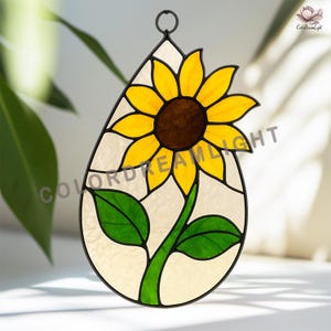 May include: Stained glass sunflower suncatcher in a teardrop shape. The sunflower has yellow petals and a brown center. Green leaves and stem are visible. The suncatcher is framed in black and has a loop for hanging. The text "COLORDREAMLIGHT" is visible.