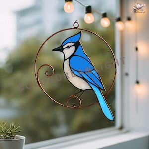 Könnte beinhalten: Ein Glasfenster-Sonnenfänger mit einem blauen Häher. Der Vogel sitzt in einem kupferfarbenen Metallkreis mit dekorativen Wirbeln. Der Vogel hat blaue, weiße und schwarze Farbtöne. Der Sonnenfänger hängt vor einem Fenster.