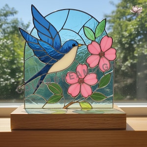 May include: Stained glass art piece featuring a blue bird with outstretched wings, a cream-colored breast, and a yellow beak. The bird is set against a blue and green background with pink flowers and green leaves. The artwork is mounted on a wooden base.
