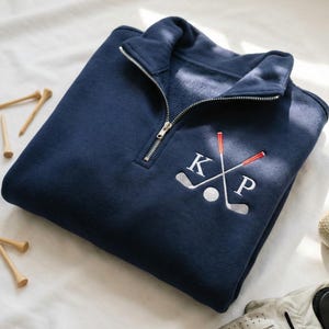 May include: Navy blue golf-themed sweatshirt with a quarter-zip collar. Features an embroidered design of crossed golf clubs, a golf ball, and the initials "KP" in white. Includes golf tees, a golf ball, and a golf glove.