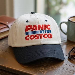 May include: A white baseball cap with a navy blue brim. The cap features the text "PANIC at the COSTCO" in red and blue lettering. The hat is sitting on a wooden surface.