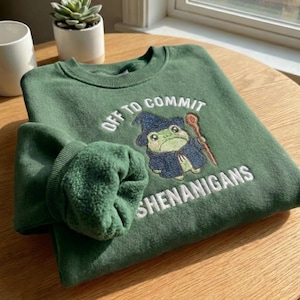 May include: A green sweatshirt with the embroidered phrase "OFF TO COMMIT SHENANIGANS" and a cartoon frog wearing a wizard hat and cloak. The sweatshirt is made of a soft material and has a crew neck.