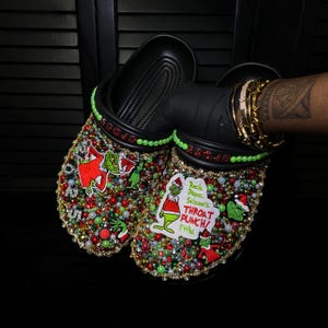 May include: Black clogs embellished with red, green, and gold beads, featuring Grinch-themed patches and text that reads "Rock Paper Scissors Throat Punch I Win". The clogs have a green trim and are decorated with various sizes of beads.