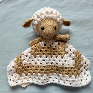 May include: A crocheted lamb security blanket with a brown and white checkered pattern. The lamb has a white fluffy head and black eyes.