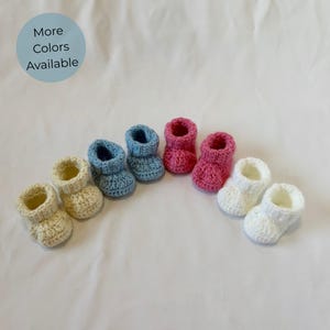 Può includere: Sei scarpine da neonato all'uncinetto in crema, azzurro, rosa e bianco. Il testo "More Colors Available" è in un cerchio azzurro. Queste morbide scarpine lavorate a maglia sono una bella idea regalo.