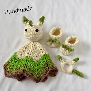 May include: A handmade crochet baby set featuring a cream-colored bunny lovey with green accents, matching booties, and a wooden ring rattle. The set includes the word "Handmade" in the image. The set is made with cream, green, and brown yarn.