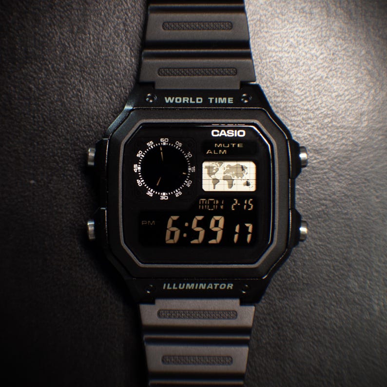 May include: Black Casio digital watch with a rectangular face. The watch displays the time, date, and world map. The watch has the text "WORLD TIME", "CASIO", and "ILLUMINATOR". The time displayed is 6:59 PM.
