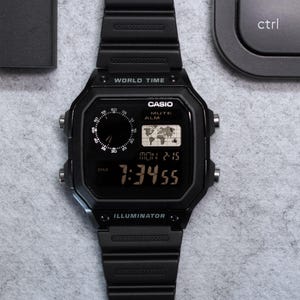 May include: A black Casio digital watch with a black band, displaying the time 7:34:55. The watch has a world map icon and the word "Casio" on the face. A black pen and a small black box are also in the image.