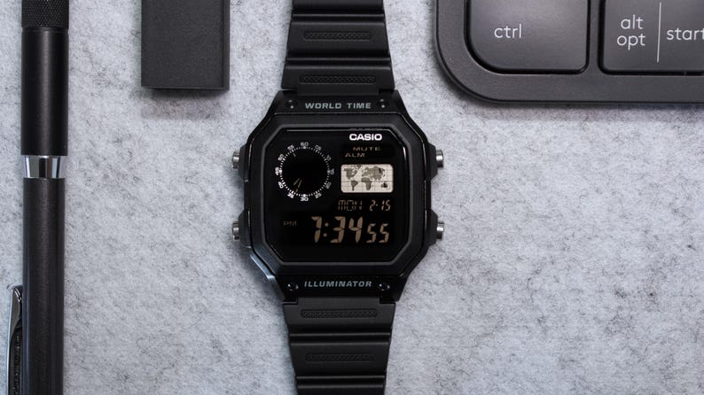 May include: A black Casio digital watch with a black band, displaying the time 7:34:55. The watch has a world map icon and the word "Casio" on the face. A black pen and a small black box are also in the image.
