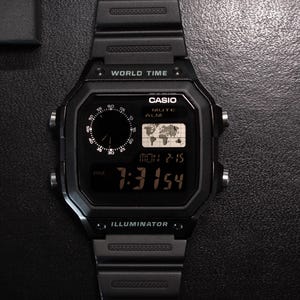 May include: Black digital Casio watch with a rectangular face, displaying the time and world map. The watch has a black band and is labeled "WORLD TIME" and "ILLUMINATOR".