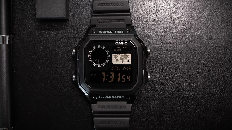 May include: Black digital Casio watch with a rectangular face, displaying the time and world map. The watch has a black band and is labeled "WORLD TIME" and "ILLUMINATOR".