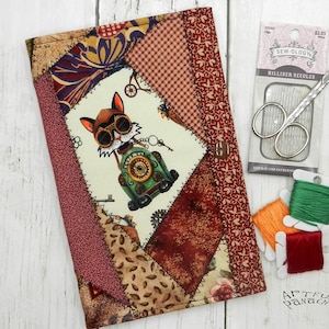 May include: A patchwork journal cover with a steampunk cat design, featuring a cat in goggles driving a green vehicle. Includes sewing supplies: needles, scissors, and spools of green, orange, and red thread. The journal cover is approximately 6 inches wide and 8 inches tall.