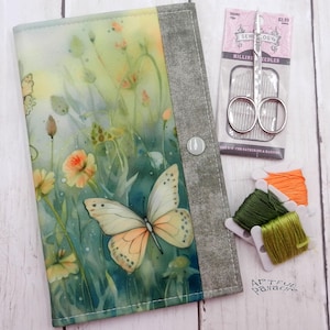 May include: A fabric notebook cover featuring a watercolor-style print of a butterfly and wildflowers, with a grey textured spine and a snap closure. Beside it are a package of Sewology millinery needles, metal scissors, and three spools of thread.