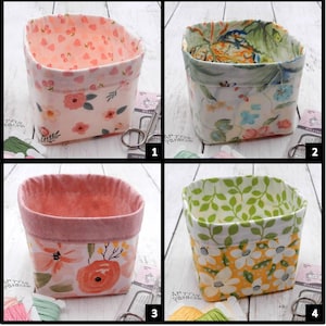 May include: Four fabric storage baskets with floral patterns. Each basket has a different design, including pink hearts, blue and pink flowers, peach flowers, and white flowers on a yellow background. The baskets are square-shaped and lined with coordinating fabric.