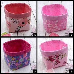 May include: Four fabric storage baskets with floral patterns. Each basket has a pink interior and a patterned exterior. The patterns include large pink flowers, small pink flowers, purple flowers, and pink flowers with green leaves. The baskets are on a white wooden surface.