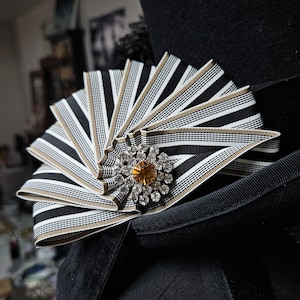 Könnte beinhalten: Schwarzer Hut mit einem dekorativen fächerförmigen Band. Das Band hat schwarz-weiße Streifen mit goldfarbenem Besatz. Eine zentrale Brosche mit einem gelben Mittelstein und klaren Kristallen sorgt für Glanz.