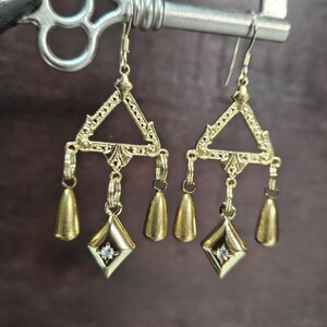 May include: A pair of gold dangle earrings with a triangular design and three teardrop-shaped charms. Each earring has a small diamond-shaped charm with a clear stone in the center.