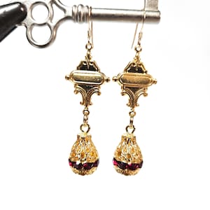 May include: Gold dangle earrings with a red stone drop. The earrings have a filigree design and a rectangular top.