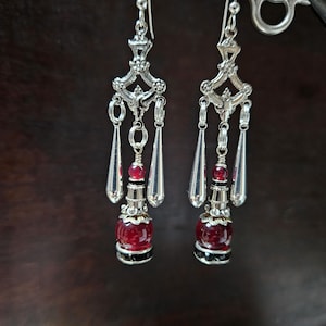 May include: Silver chandelier earrings with a decorative filigree design. Each earring features a red, round bead and dangling silver teardrop-shaped charms. The earrings have a vintage aesthetic and are suspended from a silver hook.