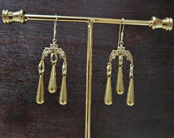 May include: A pair of gold-colored drop earrings with a decorative top and three teardrop-shaped pendants. The earrings are displayed on a gold-colored jewelry stand against a dark brown background.