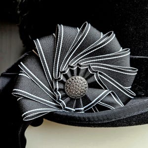 Könnte beinhalten: Eine schwarz-weiß gestreifte Schleife mit einem silbernen Knopf in der Mitte. Die Schleife ist an einem schwarzen Hut befestigt.