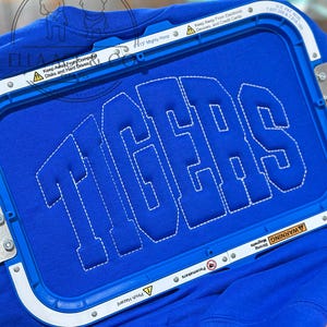 May include: A royal blue sweatshirt with the word "TIGERS" embroidered in white block letters. The sweatshirt is held in place by a blue embroidery hoop. The hoop has text that reads "Keep Away From Computer Disks and Hard Drives" and "Keep Away From Electronic Devices, and Credit Cards."