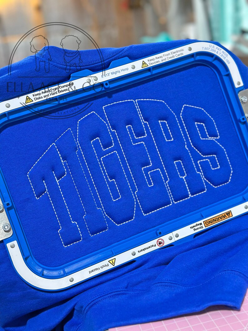 May include: A royal blue sweatshirt with the word "TIGERS" embroidered in white block letters. The sweatshirt is held in place by a blue embroidery hoop. The hoop has text that reads "Keep Away From Computer Disks and Hard Drives" and "Keep Away From Electronic Devices, and Credit Cards."