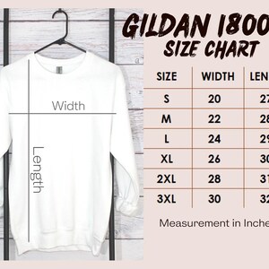 May include: A white Gildan 18000 sweatshirt hanging on a black hanger. The image includes a size chart with measurements in inches for sizes S to 3XL. The chart shows width and length measurements.