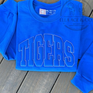May include: A royal blue sweatshirt with the word "TIGERS" stitched in white block letters. The sweatshirt has a crew neck and long sleeves. The brand name "Ella Ace & Co" is visible on a tag.
