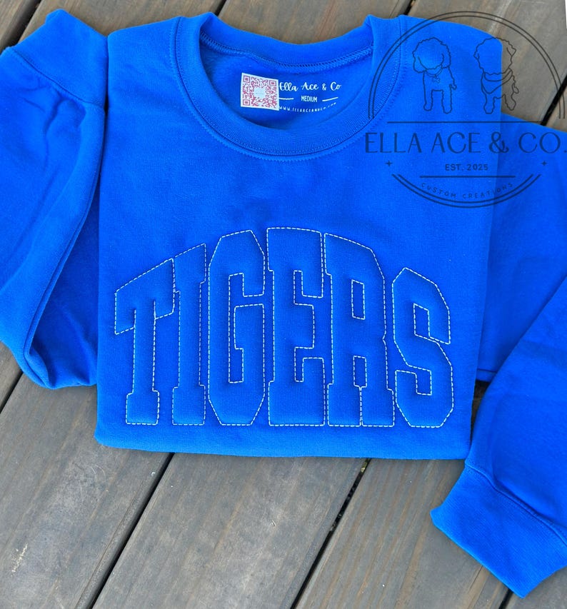 May include: A royal blue sweatshirt with the word "TIGERS" stitched in white block letters. The sweatshirt has a crew neck and long sleeves. The brand name "Ella Ace & Co" is visible on a tag.