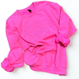 May include: A bright pink sweatshirt with the words "STROUD Tigers" embroidered on the front. The long sleeves are cuffed, and the crew neck sweatshirt is made of a soft material. The sweatshirt is displayed on a white surface.