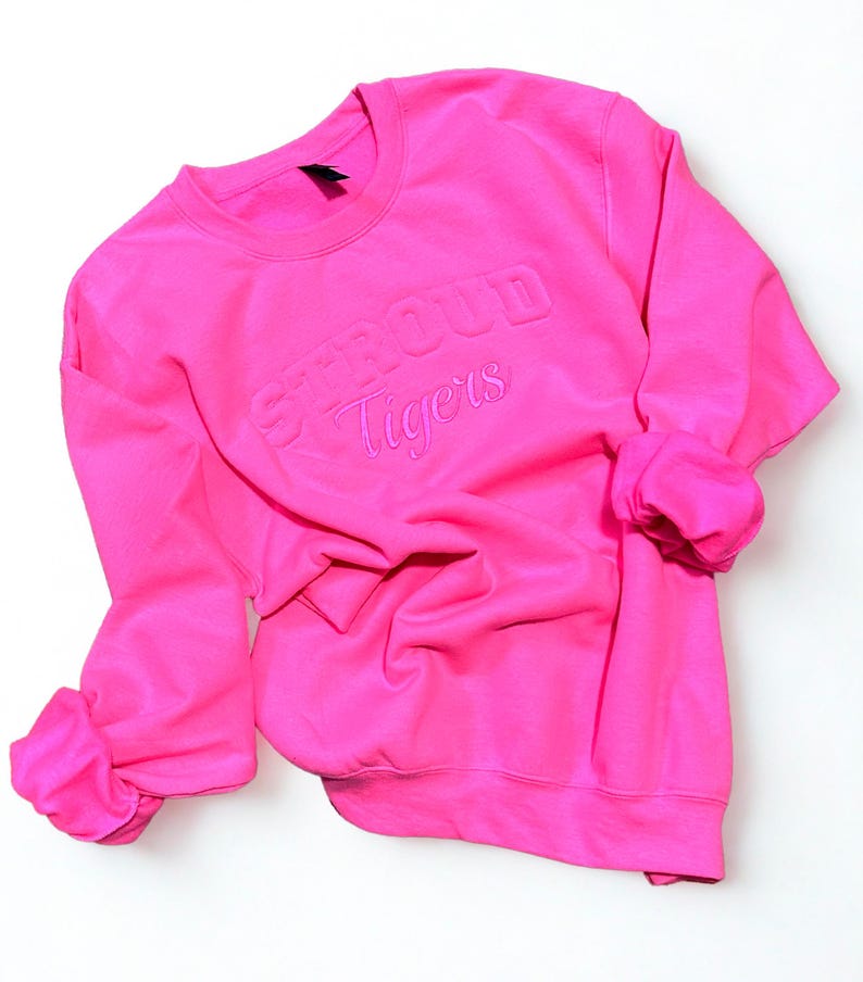 May include: A bright pink sweatshirt with the words "STROUD Tigers" embroidered on the front. The long sleeves are cuffed, and the crew neck sweatshirt is made of a soft material. The sweatshirt is displayed on a white surface.