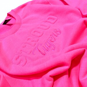 May include: Bright pink crewneck sweatshirt with the words "STROUD Tigers" embroidered in a matching pink thread. The sweatshirt is made of a soft, textured fabric and is folded, showing the neckline and part of the sleeve.