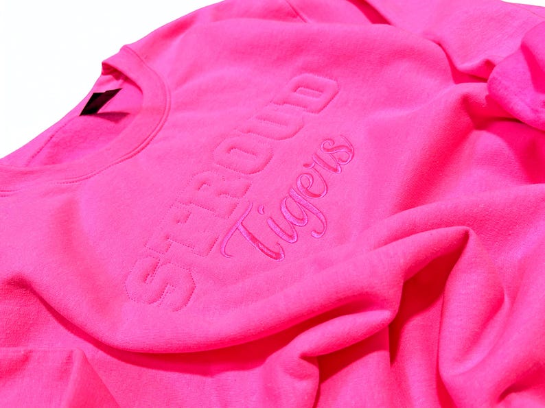 May include: Bright pink crewneck sweatshirt with the words "STROUD Tigers" embroidered in a matching pink thread. The sweatshirt is made of a soft, textured fabric and is folded, showing the neckline and part of the sleeve.