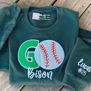 May include: Dark green sweatshirt with a baseball design. The design includes a green "G", a baseball with red stitching, and the word "Bison". The sleeve has the name "Lucas" and the number "25".
