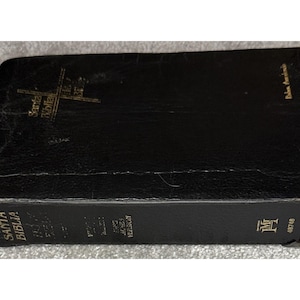 May include: A black leather-bound Bible with gold lettering. The cover features the words "Santa Biblia" and a cross design. The spine also has gold text, including the words "King James Version". The edges show some wear.