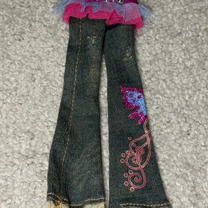 Op de afbeelding: Donkerblauwe spijkerbroek met een gerafelde zoom en een roze glitterende vlinderprint op de onderkant van de pijp. De tailleband is versierd met roze glitterstof, een lichtblauwe tule ruche en een zilverkleurige vlindervormige metalen gesp.