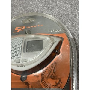 Pu&ograve; includere: Una radio per bici Sony argento e nera nella sua confezione originale. La radio ha un piccolo schermo, un altoparlante e pulsanti di controllo. La confezione riporta "SP Sports" e "Bike Radio".