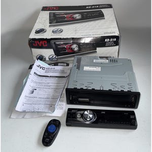 Pode incluir: Um auto-rádio JVC KD-S19 com painel frontal preto e detalhes vermelhos é exibido com sua caixa original, controle remoto e manual de instruções. A caixa mostra o nome e a imagem do produto.