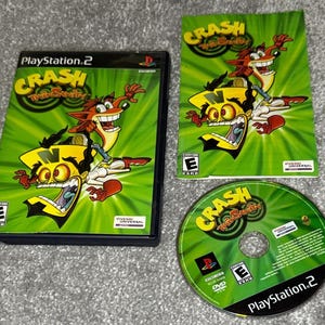 以下が含まれることがあります： PlayStation 2のゲームセットで、「Crash Twinsanity」というゲームが収録されています。 ゲームケース、カバーアート、ディスクが見え、すべて緑と黄色の配色と漫画のキャラクターグラフィックです。 ゲームは「E」レーティングです。
