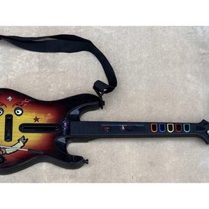Peut inclure: Une manette de jeu vidéo en forme de guitare noire et rouge avec une sangle noire. La manette présente un dessin de visage de dessin animé avec des flammes et une bannière. Les boutons de frette sont rouges, oranges, jaunes, verts, bleus et violets.