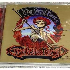 May include: A CD case featuring the album "The Very Best of Grateful Dead." The cover art showcases a skeleton figure holding a flag amidst red roses, with the band's name in a stylized font. The background is a gold color.
