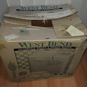 Könnte beinhalten: Beschädigter Karton für einen West Bend Automatic Bread & Dough Maker. Die Box ist beige mit grünen und schwarzen Texten und Grafiken. Die Box hat Risse und Knicke. Der Karton hat den Text "100% Whole Wheat".