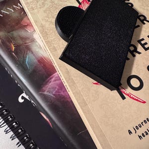 Könnte beinhalten: Ein Stapel Bücher mit einem schwarzen Buchständer obenauf. Das oberste Buch trägt den Titel "The Story We Never Wanted to Say" von Isabelle Miller. Der Buchständer hat eine strukturierte Oberfläche.