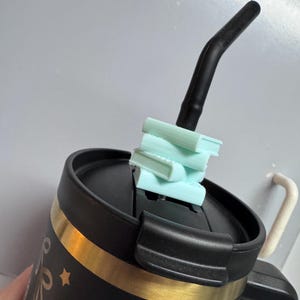 Può includere: Una tazza da viaggio nera con una fascia dorata e un motivo a stelle. La tazza ha un coperchio nero con una cannuccia che sporge. Un mucchio di oggetti di forma di libro di colore azzurro chiaro è posto sopra il coperchio.