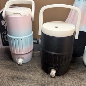 May include: Two water coolers with white handles and lids. One is black, the other is a gradient of pink and blue. Both have a white spigot at the bottom. The coolers are cylindrical with a ridged design.